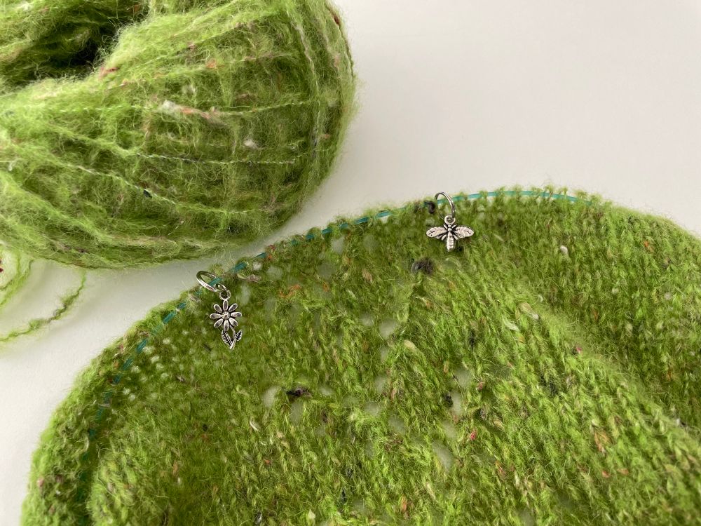 a knitting wip featuring two silver stitch markers, one bee and one flower