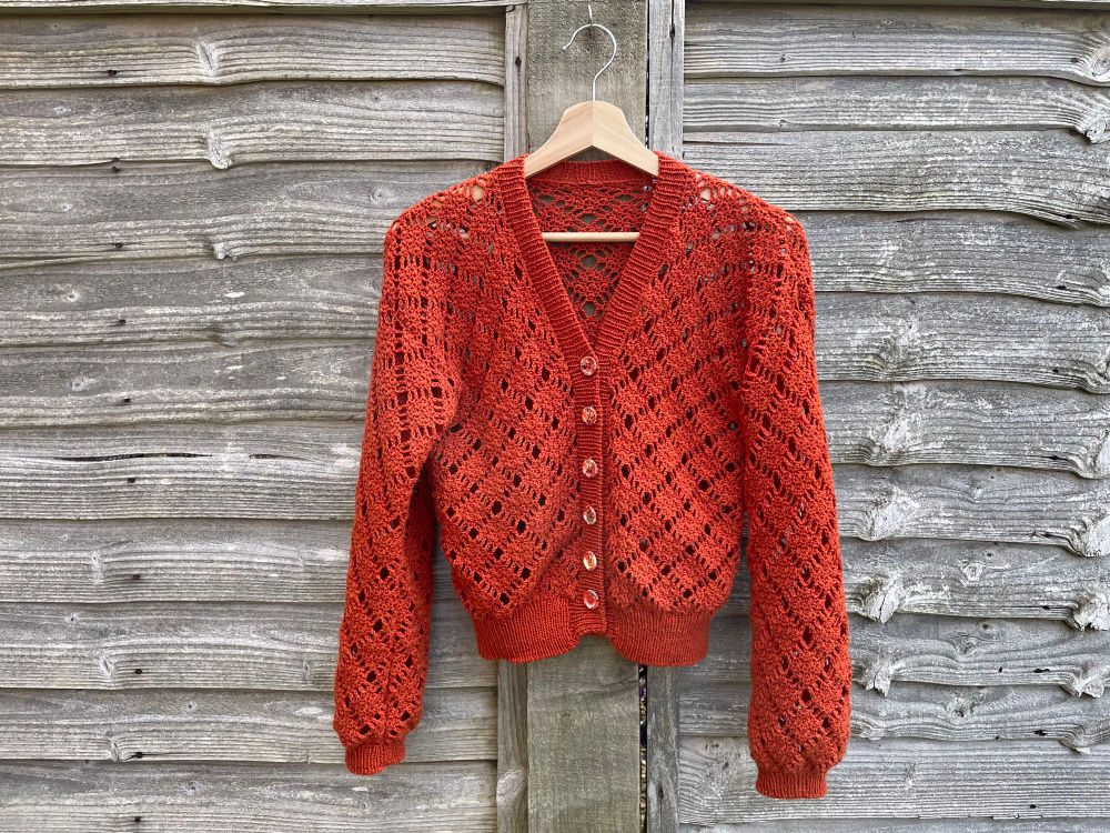 a very orange crocheted cardigan hanging against a worn fence. if you look closely you’ll see the buttons have flowers painted on them. 