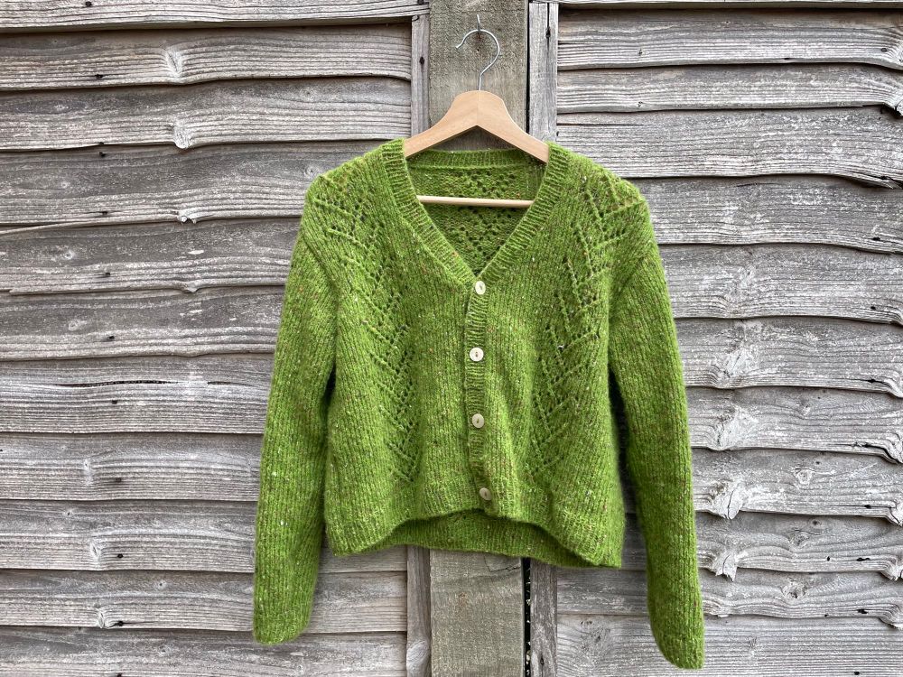 a hand-knit, grass-green cardigan hanging against a worn wooden fence. the cardigan is long-sleeved, short-bodied, made of very soft and drapey yarn, and has lace stripes climbing the front, back, and sleeves.