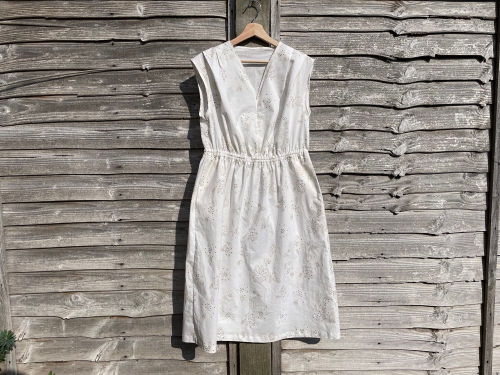 a hand-made sleeveless sundress hanging against a worn wooden fence. it’s cream with subtle printed and textured patterns of flowers and leaves.