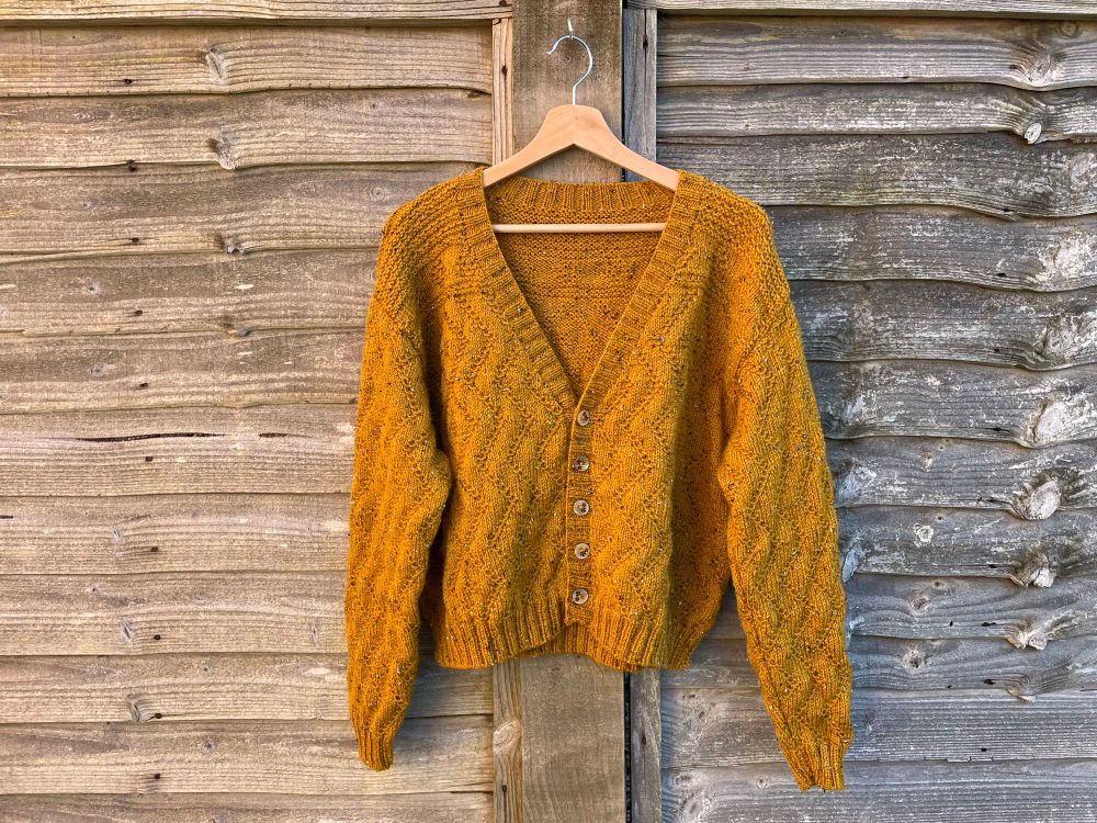 a yellow hand-knit cardigan hanging against a bare wooden fence. it has a textured pattern of diamonds and long sleeves.
