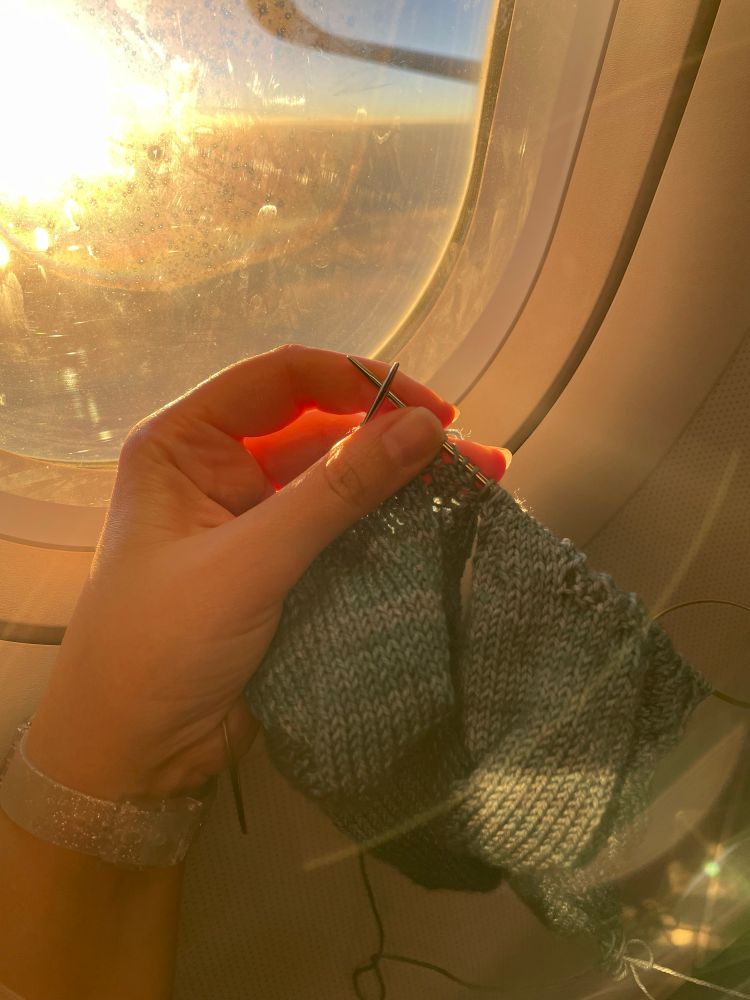 a knitting WIP held up in front of a plane window, somewhere off the coast of France iirc