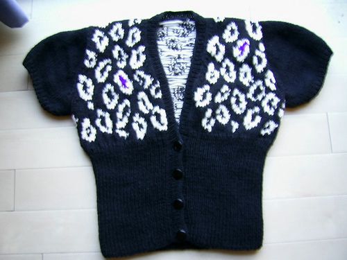 a hand-knit cardigan laid out flat. it is black with white leopard spots on the top half, and has short sleeves. 