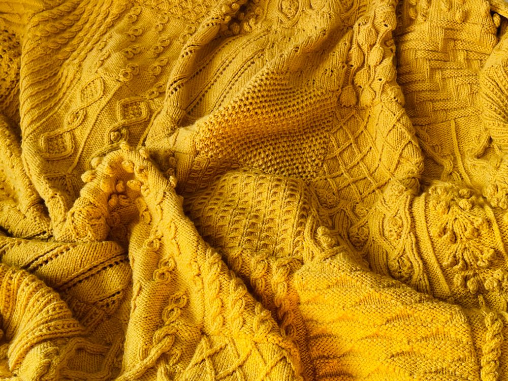 a yellow-gold knitted blanket in a variety of textured stitch patterns, scrumpled cosily and emitting a warm and joyful glow