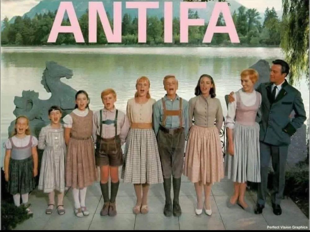 A Wes Anderson-looking photo of the Von Trapp family from the movie The Sound of Music. They are all lined up in a row on a terrace in front of a lake with beautiful trees and mountains in the background. They wear colourful pink and blue traditional Austrian clothes. Large pale pink block letters at the top say ANTIFA. image credit is Perfect Vision Graphics. 