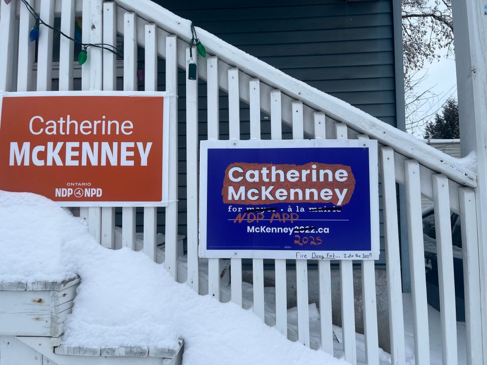 Front porch with a 2025 election sign for NDP MPP candidate Catherine McKenney, and one of McKenney’s old purple mayoral election signs, which has partly been coloured orange with a marker. “Mayor/maire” is crossed out and “NDP MPP” is written in and “McKenney2022.ca” changed to 2025. 