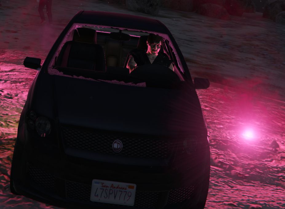 Picture from a video game. Man sitting in black car facing camera. Front window is broken. Red flare beside the car illuminating the scene.;
