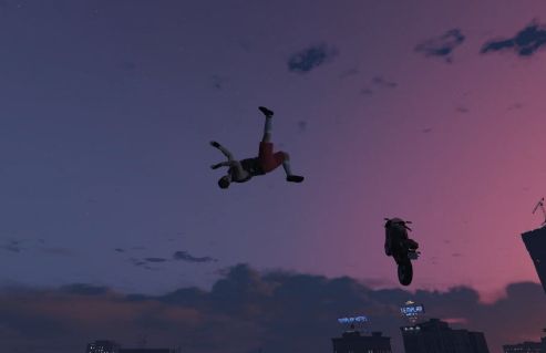 Video game character falling off of a motorcycle in mid-air, appearing to be falling. Motorcycle in the background in the air. Some buildings on the bottom left. Sunset sky with a few clouds.