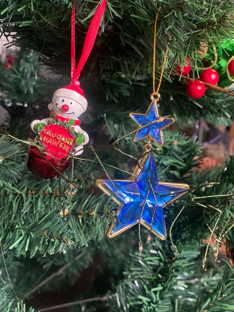 A snowman decoration with a red bell is hanging on a green Christmas tree with the words Nadolig Llawen, meaning Happy Christmas. A decoration with two blue stars is next to it. 