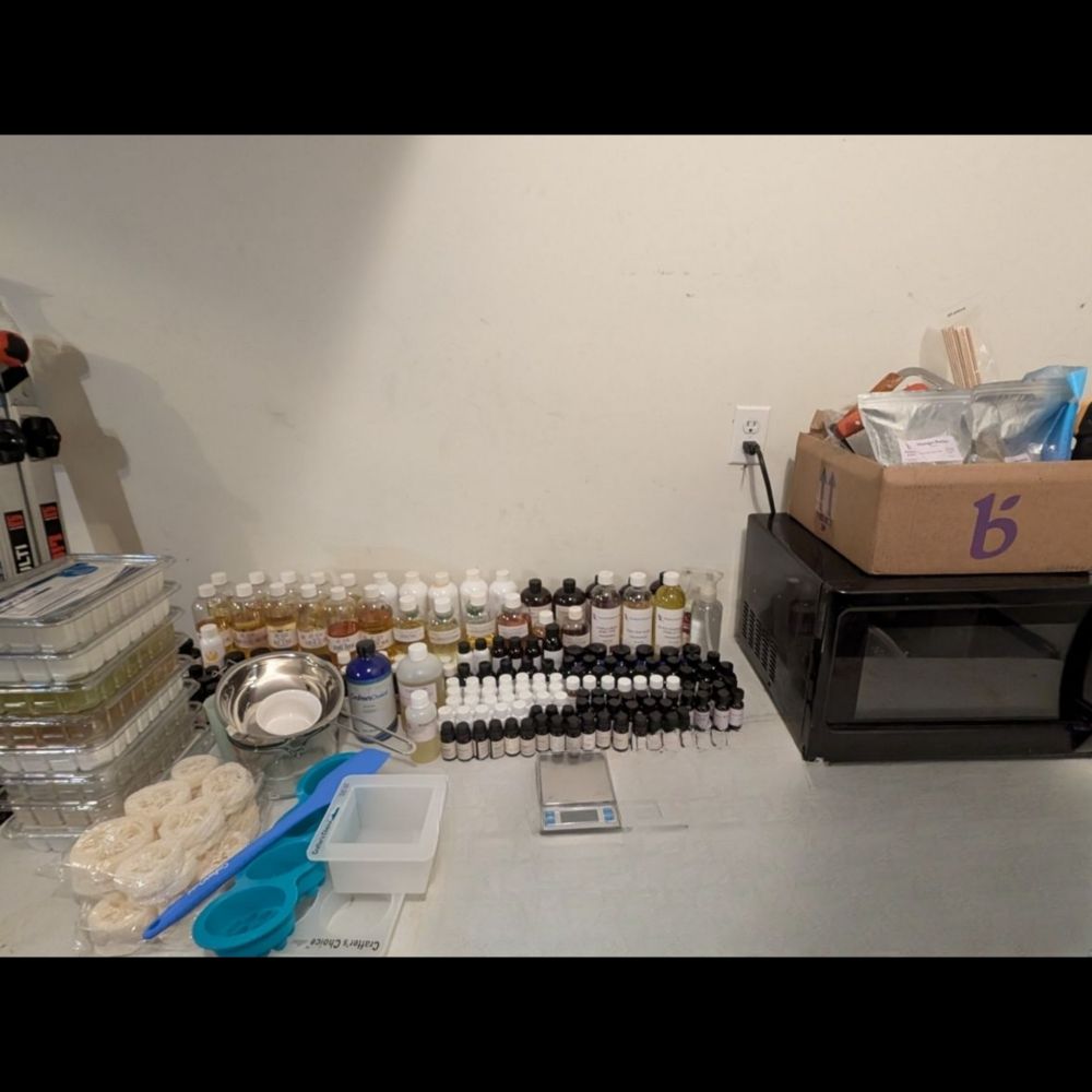 Photo of a workshop table covered in various soapmaking supplies such as soap bases, fragrance oils, molds and ingredients. A microwave oven can also be seen on the table.