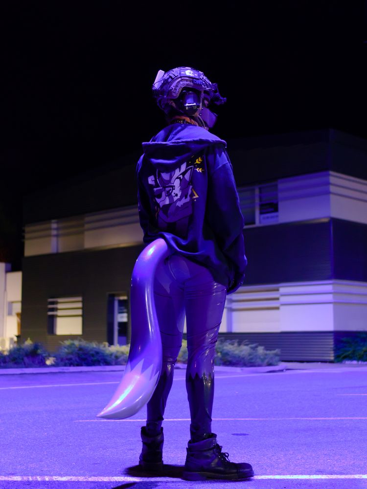 PIcture of me wearing a purple latex fox suit with inflatable tail, a hoodie with a deer furry graph in the back, a purple pup hood, and a tactical helmet. Standing in a parking lot with a purple light casting over me.