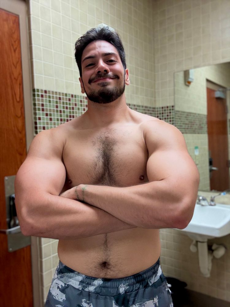 Seismic shirtless, viewed from just below the waist up, standing in a bathroom, crossing his arms to flare out his arm, chest and trap muscles.