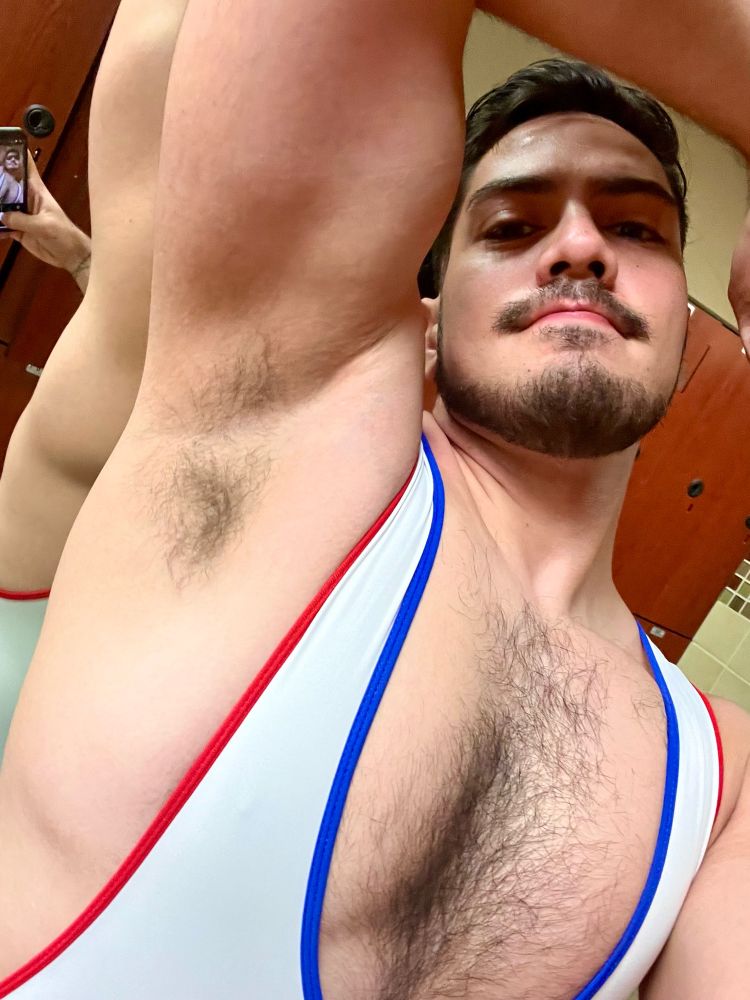 Seismic wearing a slightly low cut white singlet, with red and blue lining. He is lifting his arm above and over his head, exposing his flexed bicep and armpit to the viewer.