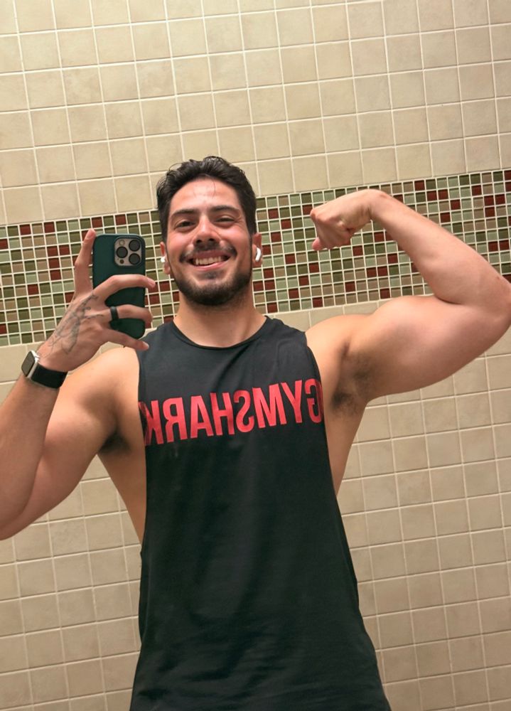 Seismic in a black Gym Shark cut off tank flexing his arm up in a bicep flex with a dopey smile on his face