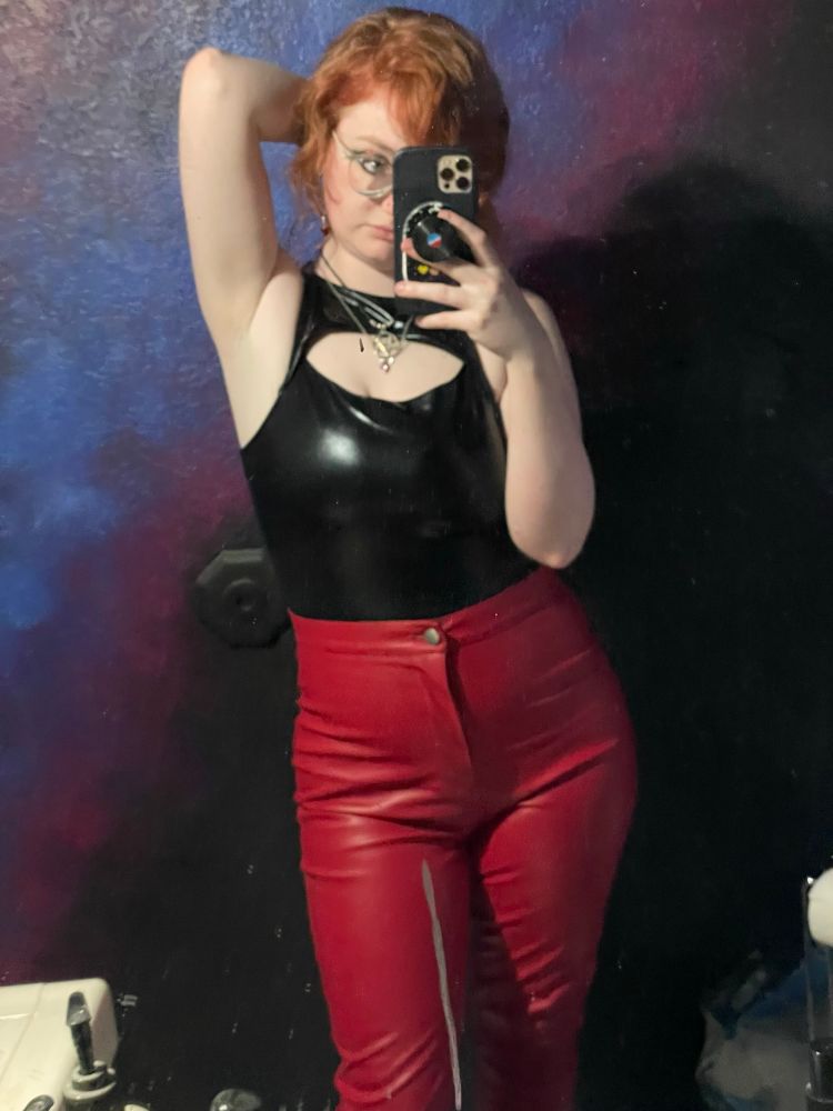 In the second photo, Helios strikes a bold pose with one arm raised and resting on his hip. He continues to wear the same black top and red pants, paired with his signature accessories. His vibrant hair stands out against the dramatic color scheme of the background. Helios looks composed and self-assured, with his phone positioned to capture his look in the mirror.