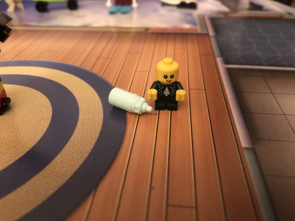 A very small Lego baby, wearing a dark green christmas jumper with a white fir tree on it. They are smiling, probably because of the pale minty coloured bottle that came with them!