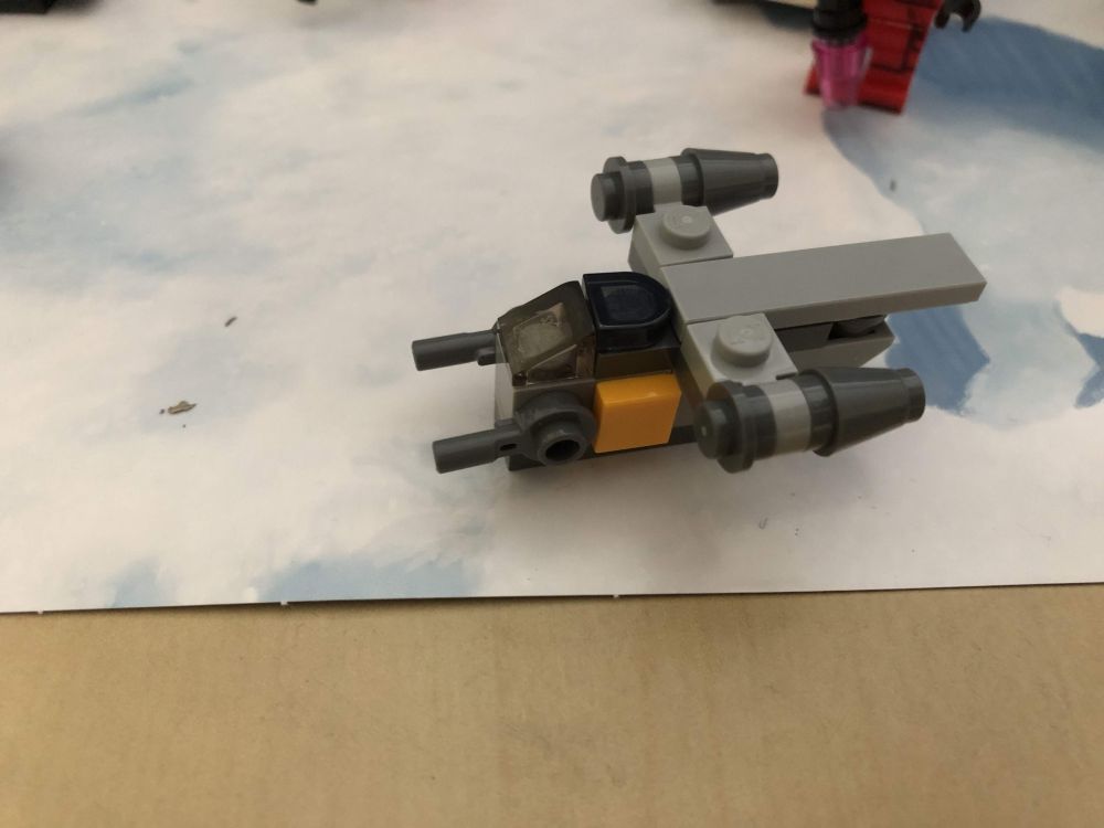This is a small version of the Razor Crest spaceship from the Mandalorian TV show, mostly grey with some dark grey engine pods along the sides and 2 small cannon at the front. There are yellow areas on either side of the ship, just behind the guns and below the cockpit.