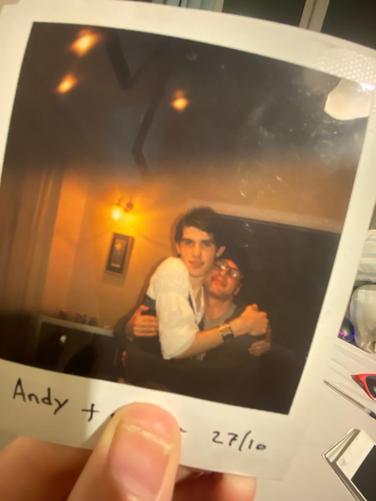 A Polaroid photo. Two teenage boys are hugging. One is wearing a T-shirt and oversized coat, the other is wearing a peasant blouse and corset.

The note on the bottom reads Andy + [redacted] 27/10
