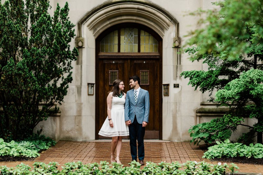 Erin on the left and her husband on the right are looking at each other. Erin is in a white dress and her husband is in a suit. They're standing in front of a wooden door in a stone building, with green trees on the sides of the frame. 