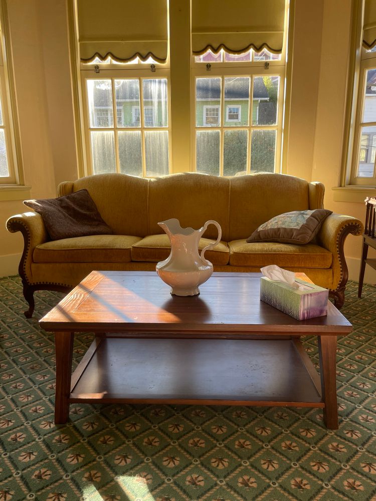 an old-fashioned parlor in yellow, faded jade, and dusty rose, last updated in the 90s. a mustard-colored sofa sits in front of a large bay window. a sunbeam illuminates a wooden coffee table on which sits a decorative white ceramic ewer.
