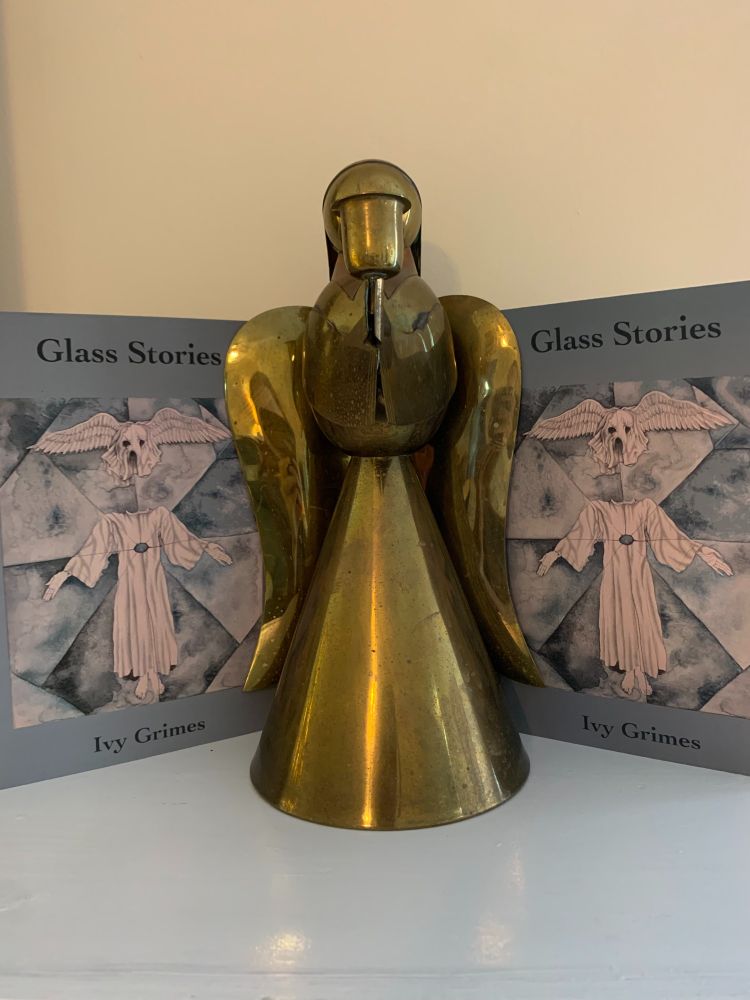 A brass angel with blue books flanking each side. The book is Glass Stories by Ivy Grimes. 