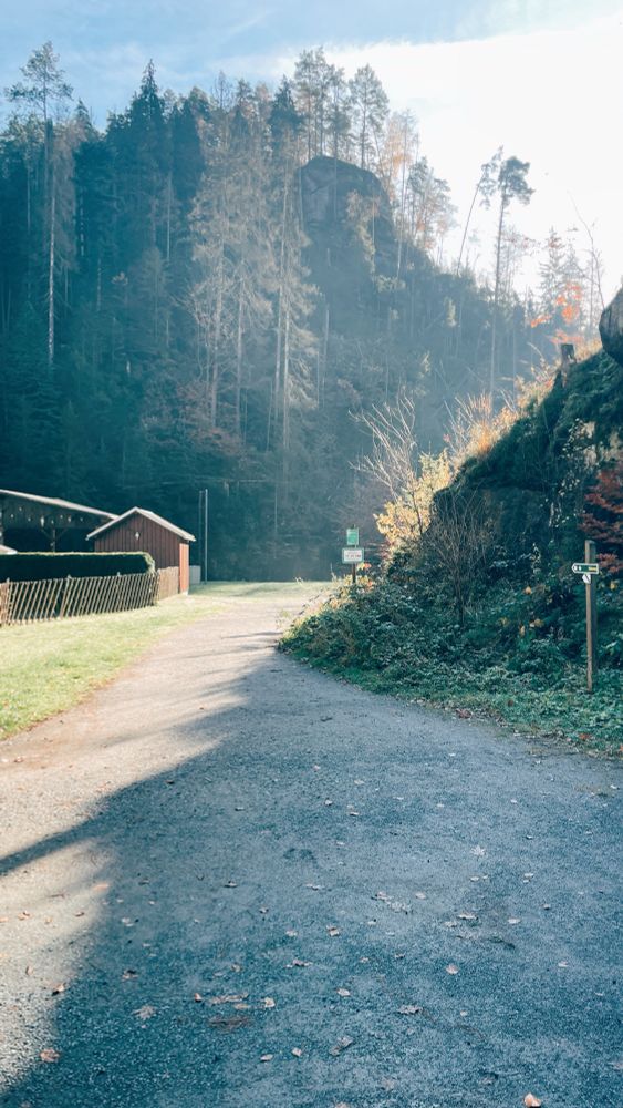 Auf der linken Seite des Bildes ist ein Wäldchen und eine Hütte zu sehen, dahinter scheint die Sonne. Auf der rechten Seite ist ein Felsvorsprung zu sehen.