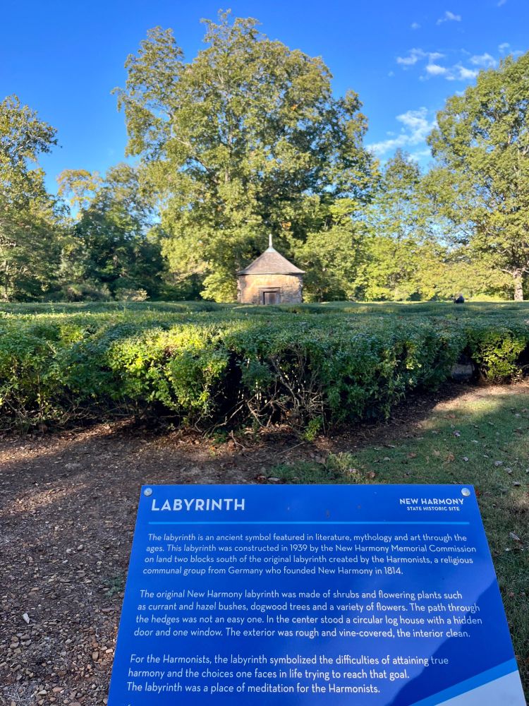 Photo of a circular stone building set in the middle of a labyrinth made of green hedges. In the foreground is a blue sign with information on labyrinths and their significance in New Harmony’s history.
