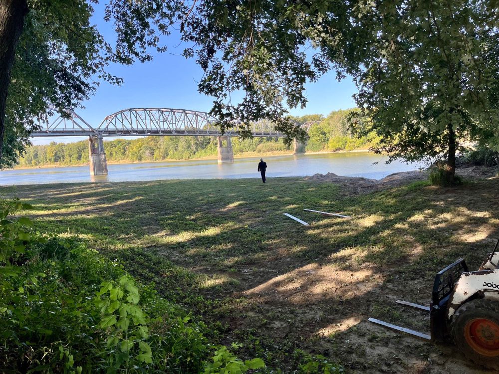 Photo of a person walking on the shore of a river. Spanning the river is an older metal bridge.