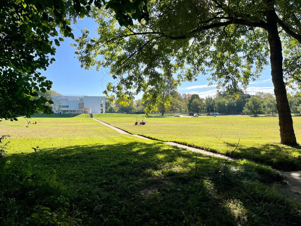 Photo of a white modernist building set in a hill above a green lawn. In the foreground is a golf cart and trees.