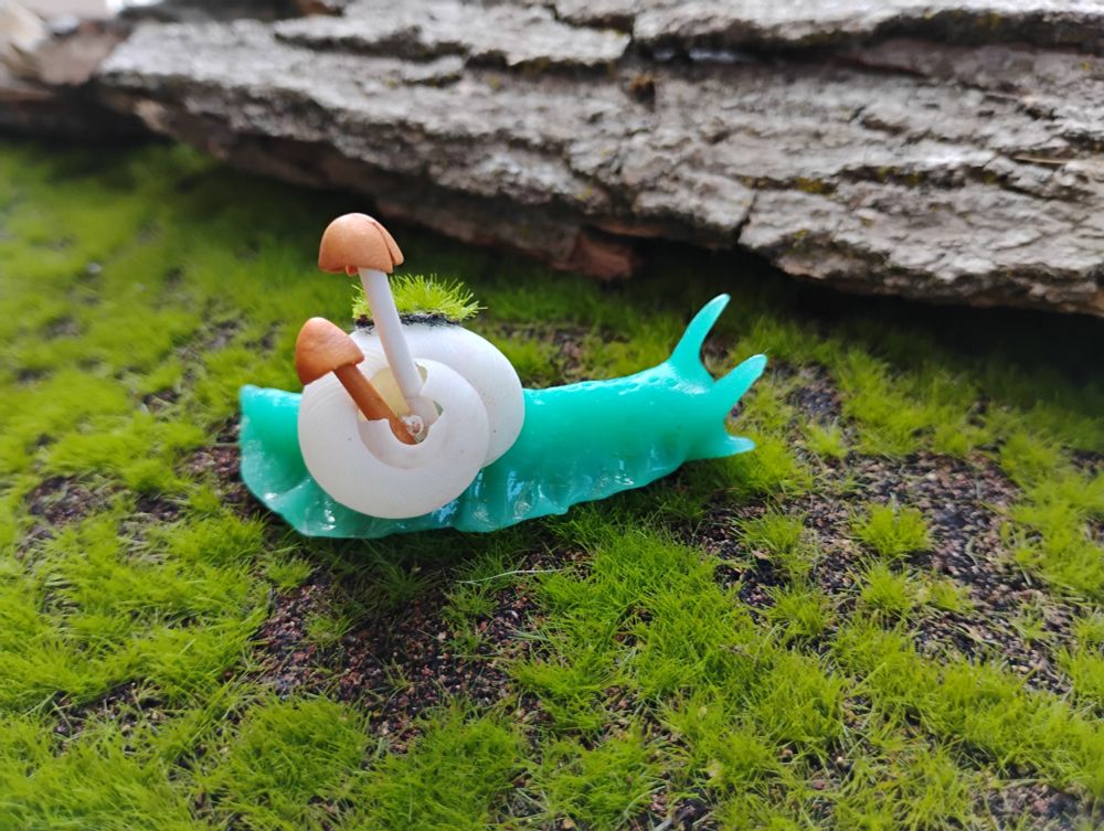 A photo of a snail brooch on a grassy background. The snail is hand sculpted out of teal colored semi-translucent polymer clay. The shell is real, it is white and has a crack at it's center from which two tiny tan and white mushrooms are sprouting. The mushrooms are also made of polymer clay.   