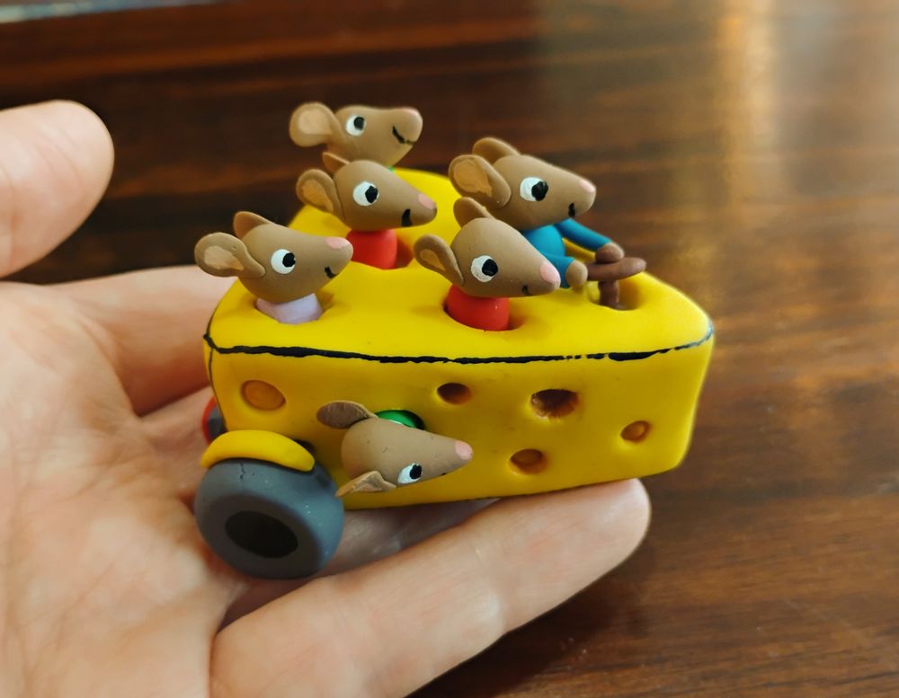 A photo of a sculpture made of colored Sculpey Clay. The sculpture is based on the Richard Scarry drawing of 6 mice riding in a car made out of a triangle block of yellow Swiss cheese. The little happy mice poke their heads out of the holes in the cheese. It's really cute imo, he popped off when he drew this one. The entire sculpture is about three inches long and fits in the artist's hand. 