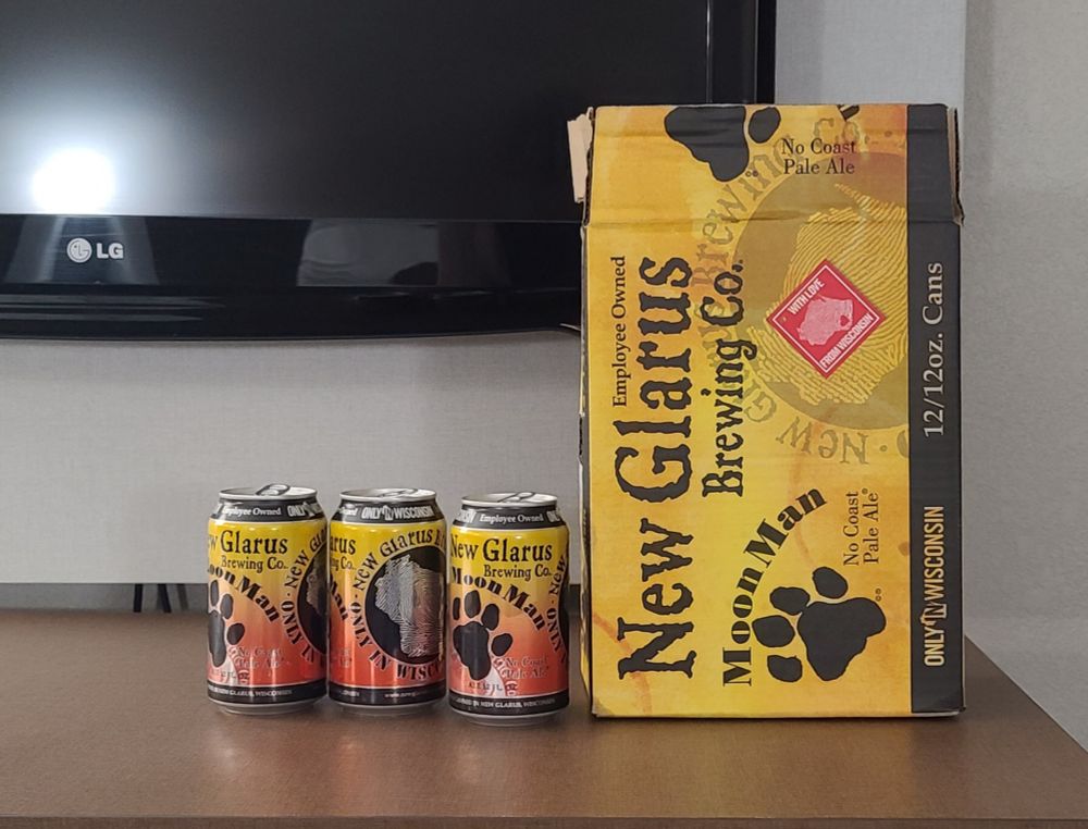 An open twelve-pack box of New Glarus Moon Man ale, with three open cans from the box next to it, on a countertop in front of an LG TV. 