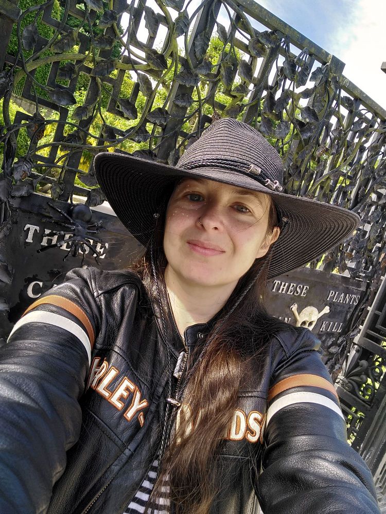 Ally, a white skinned person with long brown hair, is wearing a black leather jacket, a large black wide-brimmed hat, and taking a selfie in  front of ornate black iron gates which read "these plants can kill"