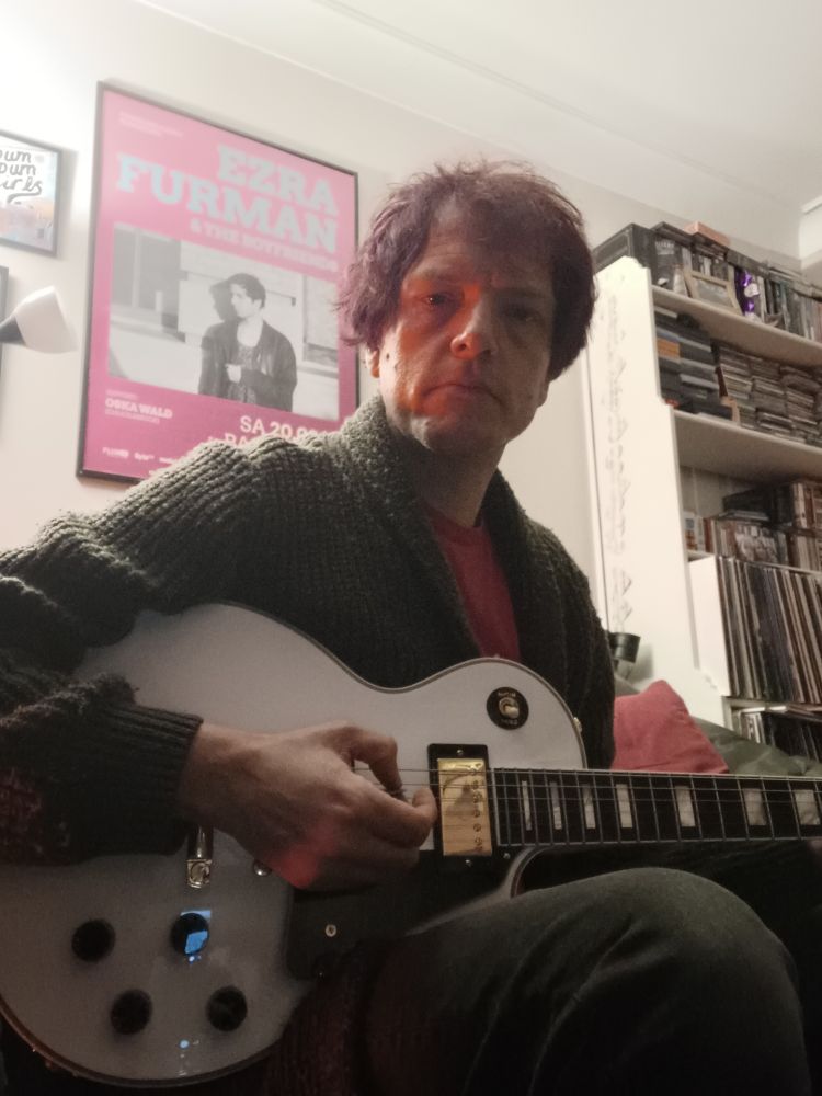 Colour photo of a very stoned 45 year old man with dyed purple hair, wearing black jeans and a red t-shirt slightly visible beneath a massive sage green Cobain style cardy. Overall look of tragic-middle-aged-grungeboi disaster offset by his guitar, an alpine white Les Paul Custom of the James Dean Bradfield variety, an axe of class and beauty with an iconic aesthetic of such force that even Stu can't make it look scruffy.