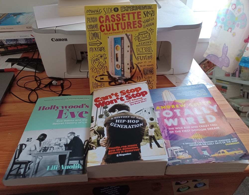 Three books laid face up on a messy desk with a fourth standing upright at the back, propped up against a printer. That book is Cassette Culture by Bentelli. Then l-r across the front Hollywood's Eve by Lili Anolik, Can't Stop, Won't Stop by Jeff Chang, and Totally Wiredk the wild rise and crazy fall of the first dotcom bubblye by Andrew Smith.