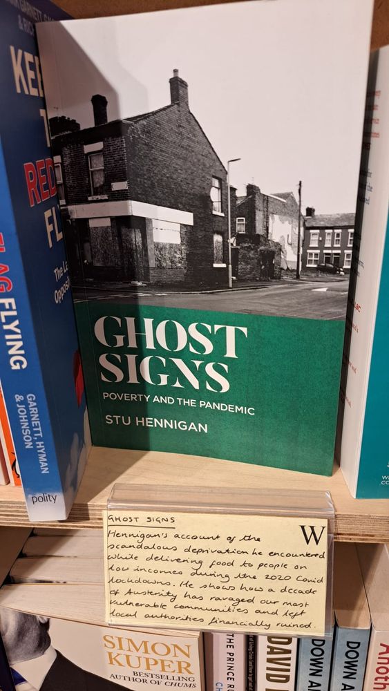 A front on colour photo of a paperback copy of the book Ghost Signs by Stu Hennigan, on display in Waterstones Tottenham Court Road with a handwritten postcard of recommendation pinned to the shelf below it.