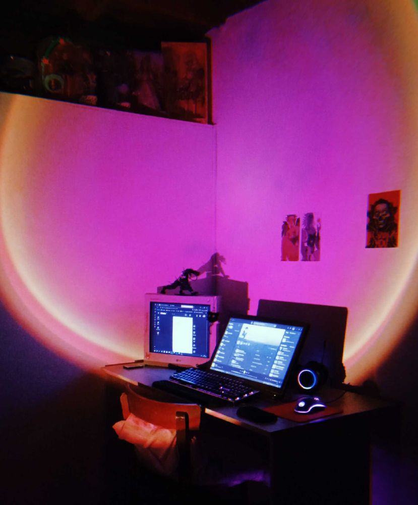 Picture of my studio set up. A white desk with two monitors on it. A display Huion tablet and a crt 2006 desktop monitor, there is a figure on top of it and three posters on the white wall. On the top of a room divider wall there are dolls boxes. The room is lit with a sunset lamp.