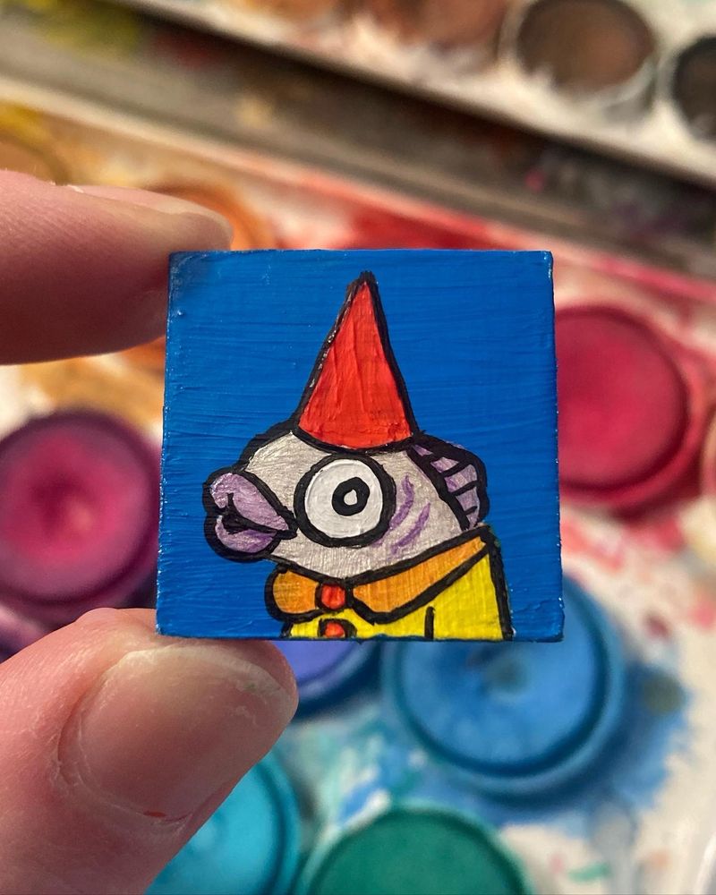 Scrabble tile painted with acrylics. Features a cartoon fish dressed like a gnome. Blue background, silver anthropomorphic fish with yellow shirt and red hat. Shirt has orange collar and red buttons. The fish has purple lips and looks surprised.