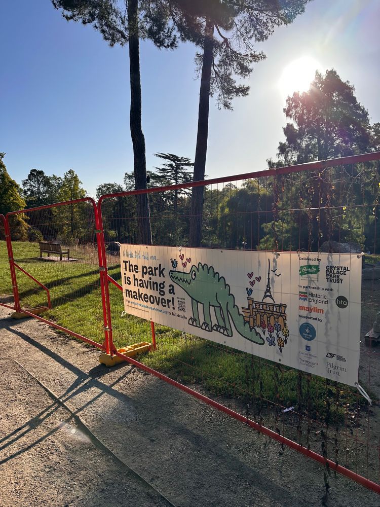 Colour photo. Orange fencing on a sunny morning, a banner with a cartoon dinosaur and company/council logos proclaims “The park is having a makeover”
