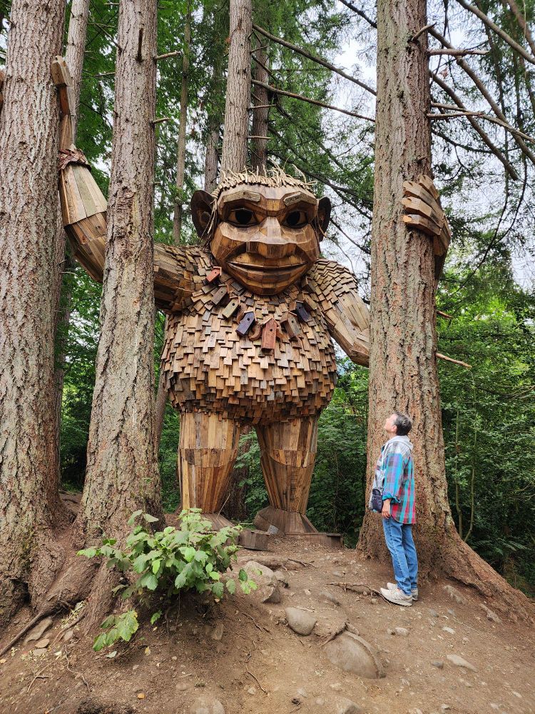 Gigantic wooden sculpture of a troll between four large trees. His hands wrapped around two of them. My lovely wife is standing next to the troll looking up at him and is dwarved by the sculpture. He is wearing a necklace of birdhouses to interact even more with nature.