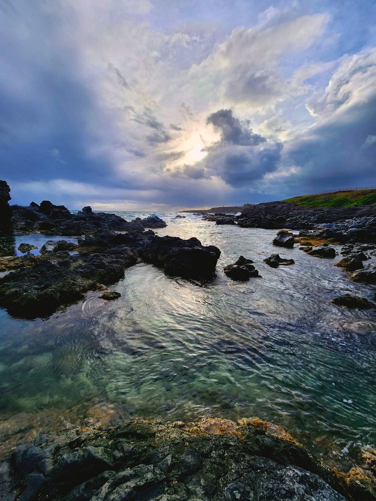 Ocean and rocky land in a shallow angle photo. Water is green and reflective because of the low sun. Ocean is cut up and dotted by lava rocks and shoreline. Sky is somewhat cloudy with deep blue colors.