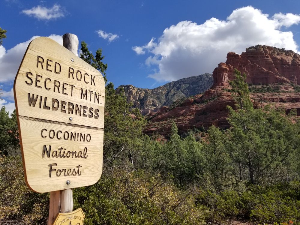 Shot of Sedona red rock country with large outcroppings and a covering of trees nearby. There is a sign that says "Red Rock Secret Mtn. Wilderness - Coconino National Forest"