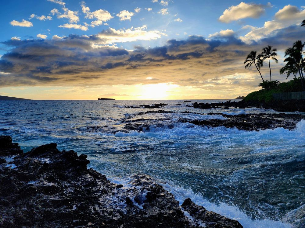 Sunset photo of crashing ocean against rocks. Water is many shades of blue, as is the sky. Much of the water is white from waves crashing and the white is cast in blue also.