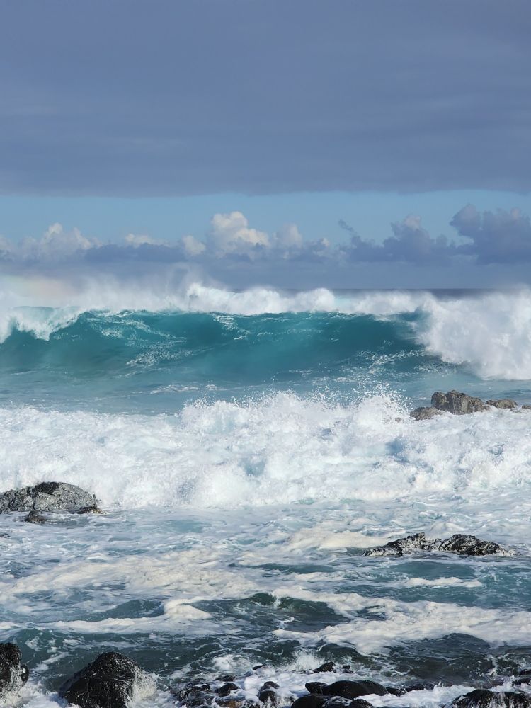 Photo from.shore of several rows of incoming waves breaking and foamy. Green transparent face on biggest wave. Dark and puffy clouds low in priorizing hard to distinguish from waves.