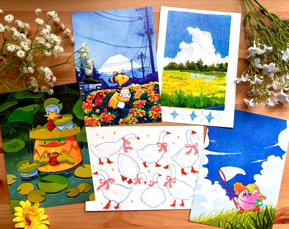 A photo of 5 different art prints with my artworks, laying on a wooden desk, surrounded by flowers.