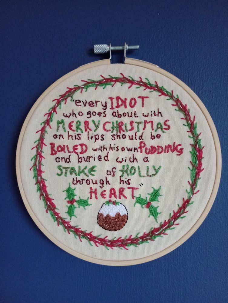 An embroidery hoop is hanging on a blue wall. Words are embroidered on a cream background in red, green and black text within a red and green wreath above a Christmas pudding and two sprigs of holly. The text reads " every idiot who goes about with Merry Christmas on his lips should be boiled with his own pudding and buried with a stake of holly through his heart". It is from A Christmas Carol by Charles Dickens.