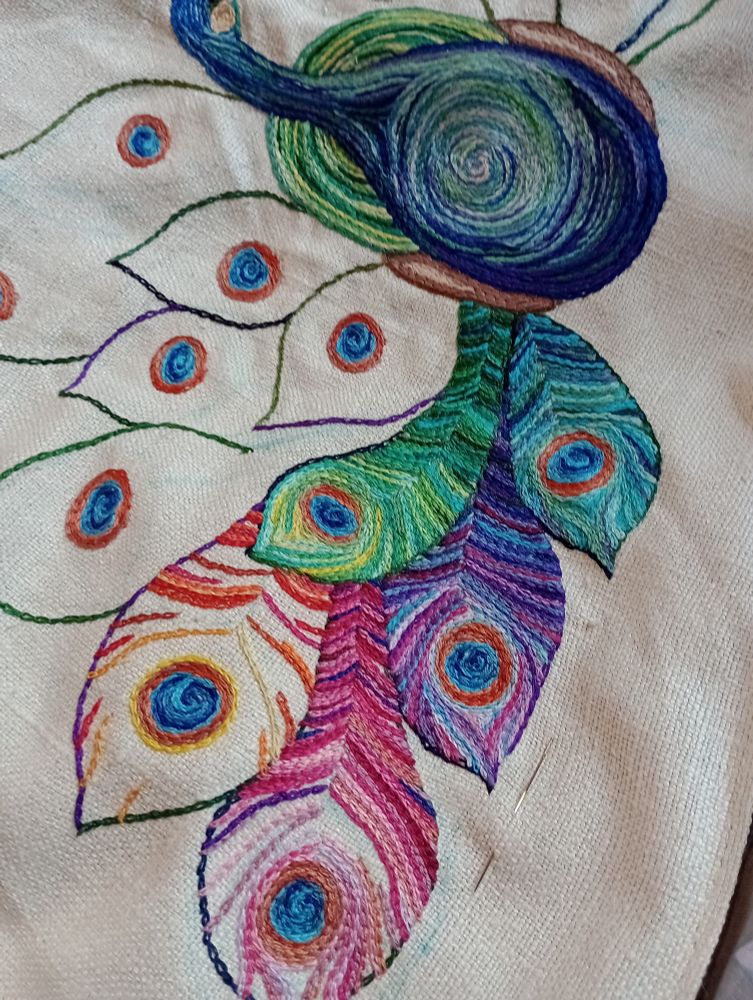 The feather shapes surrounding a peacock body filled in with varying shades of embroidery in lines of chain stitch. Green, blue and purple feathers are completed and pink and orange feathers are in progress.