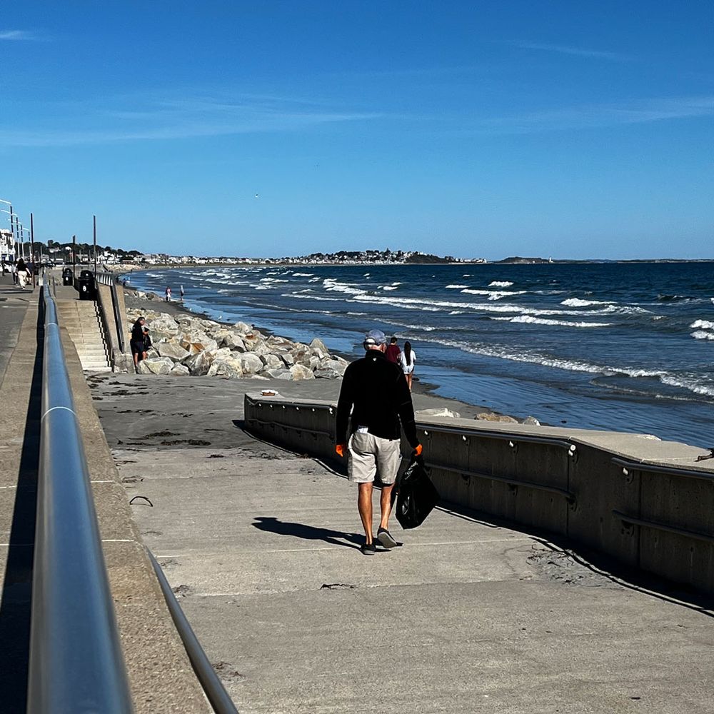 A few volunteers during a Nantasket Beach cleanup. A man in the foreground is carrying a partially full trash bag while walking down ramp to the beach, which has lots of waves.