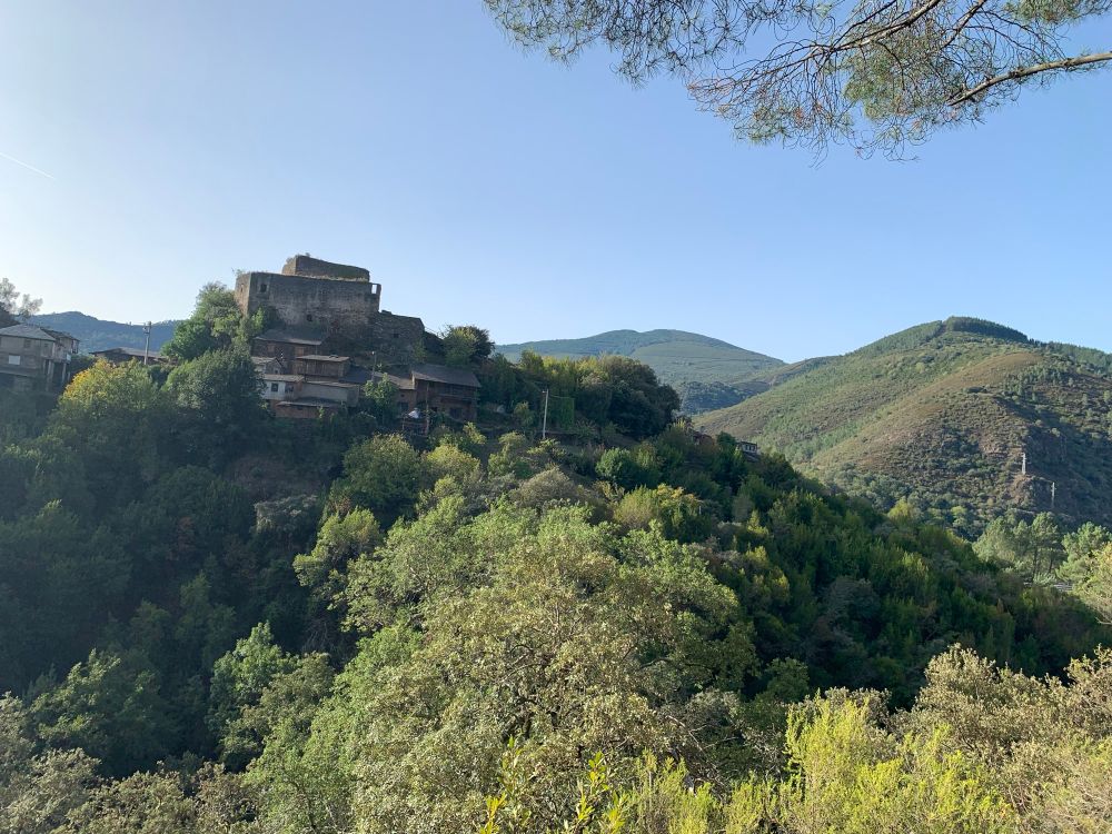 A view of a hill with a medieval stone structure at the top, surrounded by trees and greenery. In the background, rolling hills and a clear blue sky are visible.