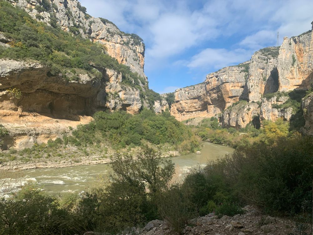 A scenic view of a rocky gorge with towering cliffs, lush greenery at the base, and a river flowing through the valley. The sky is partly cloudy, creating a serene outdoor landscape.
